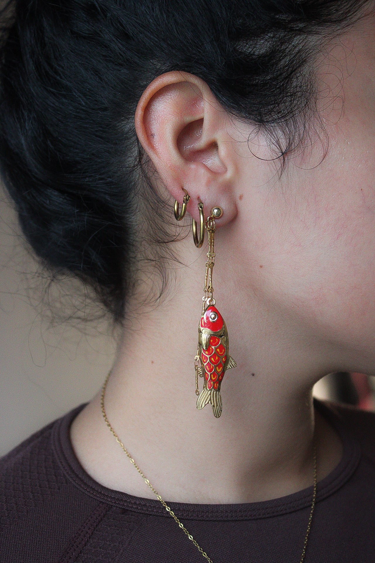A single dangling fish-shaped earring in gold-tone metal with red and orange enamel detailing, photographed on a blue grid-patterned background. Pantallas artesanales de Marveyra, hechas a mano en Puerto Rico con materiales resistentes. Este diseño de joyería alternativa y unisex cuenta con elementos removibles que permiten personalizar la pieza o utilizar los colgantes por separado como collar. Accesorios originales creados para quienes buscan destacar con un estilo diferente. Pantallas de peces.