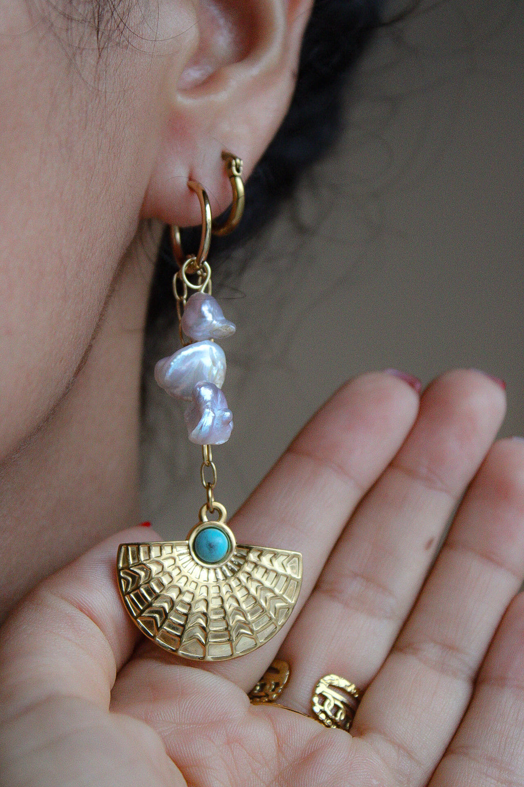 A close-up of a person's ear and hand wearing a gold hoop earring with dangling irregular white pearl-like beads and a gold fan-shaped charm featuring a small turquoise stone. Pantallas artesanales de Marveyra, hechas a mano en Puerto Rico con materiales resistentes. Este diseño de joyería alternativa y unisex cuenta con elementos removibles que permiten personalizar la pieza o utilizar los colgantes por separado como collar. Accesorios originales creados para quienes buscan destacar con un estilo diferente