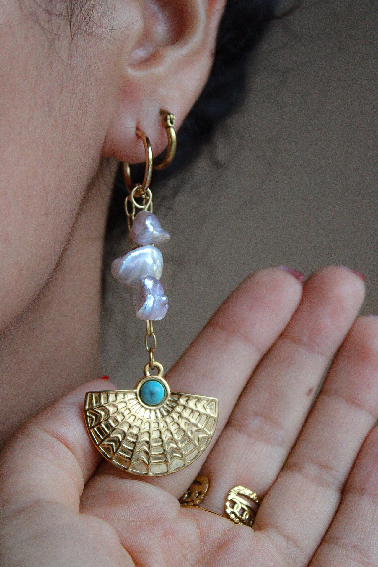 A close-up of a person's ear and hand wearing a gold hoop earring with dangling irregular white pearl-like beads and a gold fan-shaped charm featuring a small turquoise stone. Pantallas artesanales de Marveyra, hechas a mano en Puerto Rico con materiales resistentes. Este diseño de joyería alternativa y unisex cuenta con elementos removibles que permiten personalizar la pieza o utilizar los colgantes por separado como collar. Accesorios originales creados para quienes buscan destacar con un estilo diferente