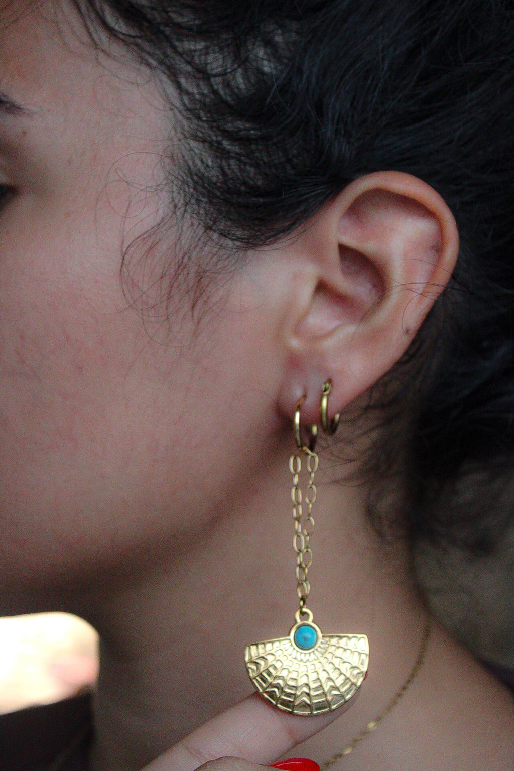A close-up of a person's ear wearing a gold dangle earring with a textured fan-shaped drop and a small round turquoise blue stone at the top.Pantallas artesanales de Marveyra, hechas a mano en Puerto Rico con materiales resistentes. Este diseño de joyería alternativa y unisex cuenta con elementos removibles que permiten personalizar la pieza o utilizar los colgantes por separado como collar. Accesorios originales creados para quienes buscan destacar con un estilo diferente.