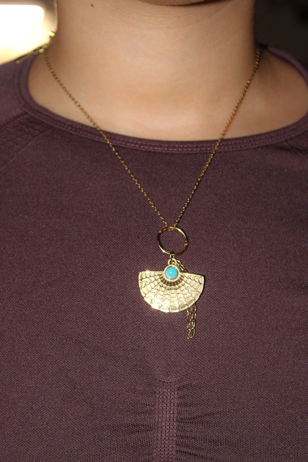 A gold pendant necklace worn by a person, featuring a textured fan-shaped pendant with a small turquoise blue stone at the center, set against a dark plum top.Pantallas artesanales de Marveyra, hechas a mano en Puerto Rico con materiales resistentes. Este diseño de joyería alternativa y unisex cuenta con elementos removibles que permiten personalizar la pieza o utilizar los colgantes por separado como collar. Accesorios originales creados para quienes buscan destacar con un estilo diferente.
