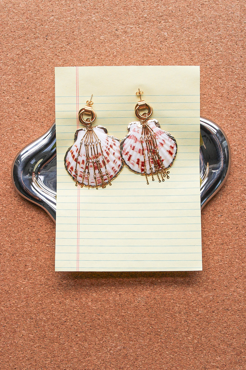 A pair of handmade shell earrings with gold-tone rings and dangling fringe, displayed on a piece of lined paper over a silver tray against a cork background. A scallop shell pendant with red and white stripes, decorated with gold-toned chains and a gold clasp, displayed on a blue denim fabric background. Par de pantallas de concha caracol con cadenas colgantes doradas, grades y unicas, estilo llamativo. Pantallas playeras y tropicales a la moda.