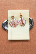 A pair of handmade shell earrings with gold-tone rings and dangling fringe, displayed on a piece of lined paper over a silver tray against a cork background. A scallop shell pendant with red and white stripes, decorated with gold-toned chains and a gold clasp, displayed on a blue denim fabric background. Par de pantallas de concha caracol con cadenas colgantes doradas, grades y unicas, estilo llamativo. Pantallas playeras y tropicales a la moda.