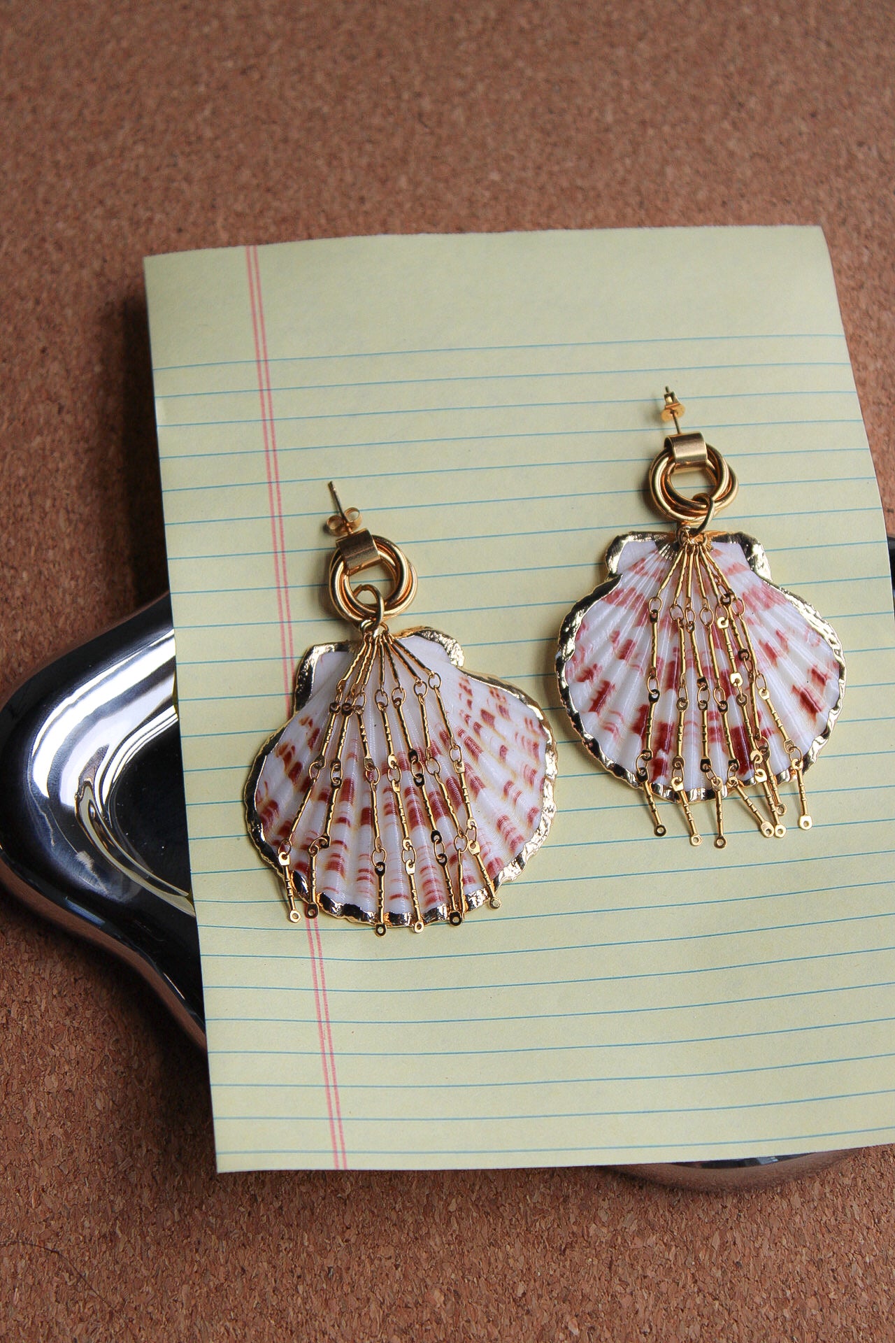 A pair of dangling shell-shaped earrings with gold-tone hooks and wireframe detailing, displayed on a sheet of lined paper over a brown textured surface. A scallop shell pendant with red and white stripes, decorated with gold-toned chains and a gold clasp, displayed on a blue denim fabric background. Par de pantallas de concha caracol con cadenas colgantes doradas, grades y unicas, estilo llamativo. Pantallas playeras y tropicales a la moda.