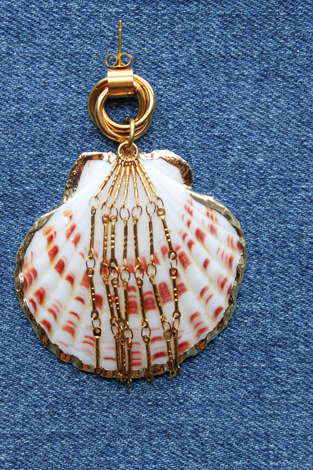A scallop shell pendant with red and white stripes, decorated with gold-toned chains and a gold clasp, displayed on a blue denim fabric background. Par de pantallas de concha caracol con cadenas colgantes doradas, grades y unicas, estilo llamativo. Pantallas playeras y tropicales a la moda.
