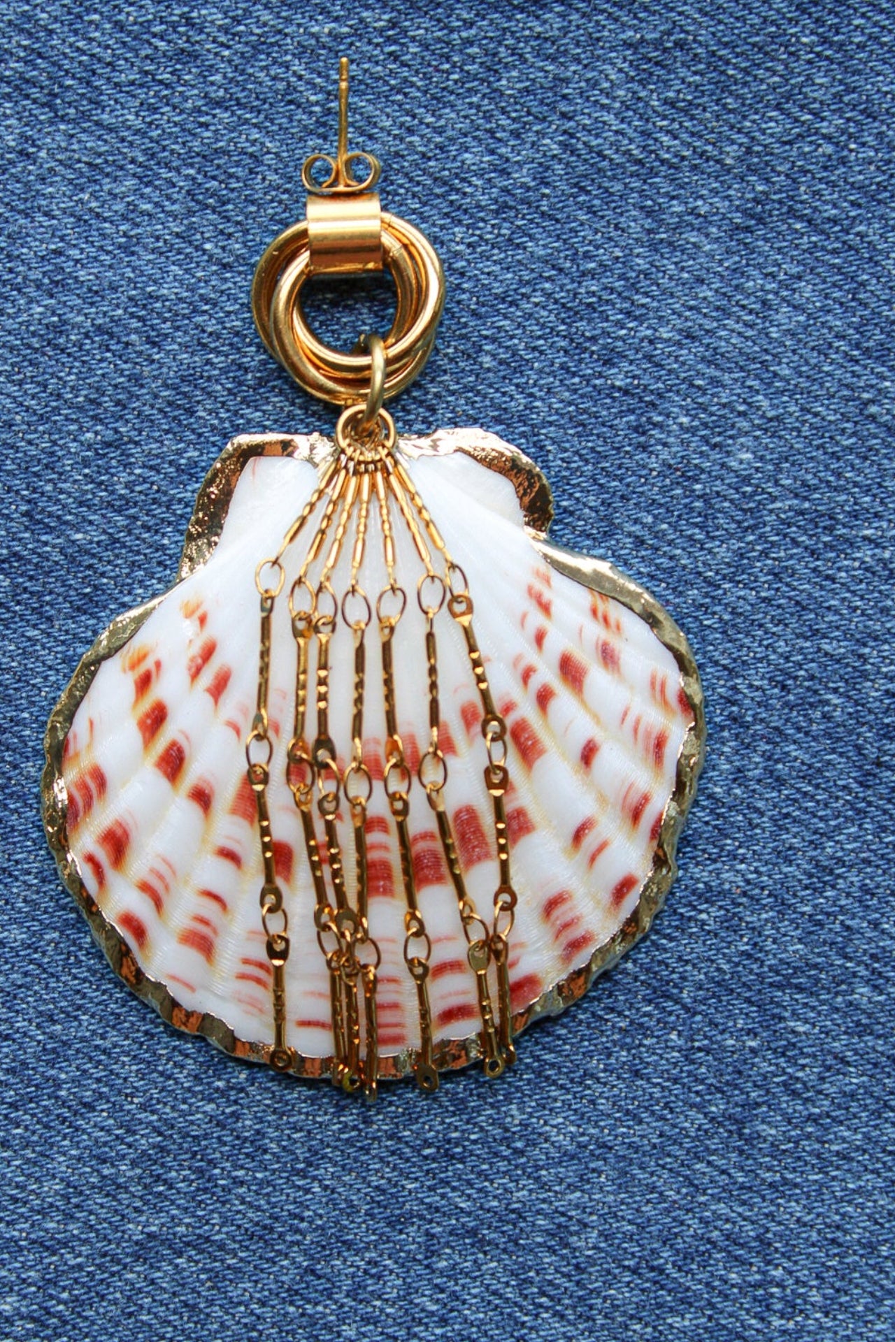 A scallop shell pendant with red and white stripes, decorated with gold-toned chains and a gold clasp, displayed on a blue denim fabric background. Par de pantallas de concha caracol con cadenas colgantes doradas, grades y unicas, estilo llamativo. Pantallas playeras y tropicales a la moda.