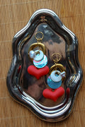 A pair of handmade dangle earrings displayed on a reflective silver tray over a woven bamboo mat. The earrings feature gold-tone rings, white pearl-like beads, iridescent blue beads, and red heart charms. Un par de pantallas colgantes artesanales sobre una bandeja plateada reflectante y un mantel de bambú tejido. El diseño incluye aros dorados, cuentas tipo perla blancas, cuentas azules iridiscentes y dijes de corazones rojos.