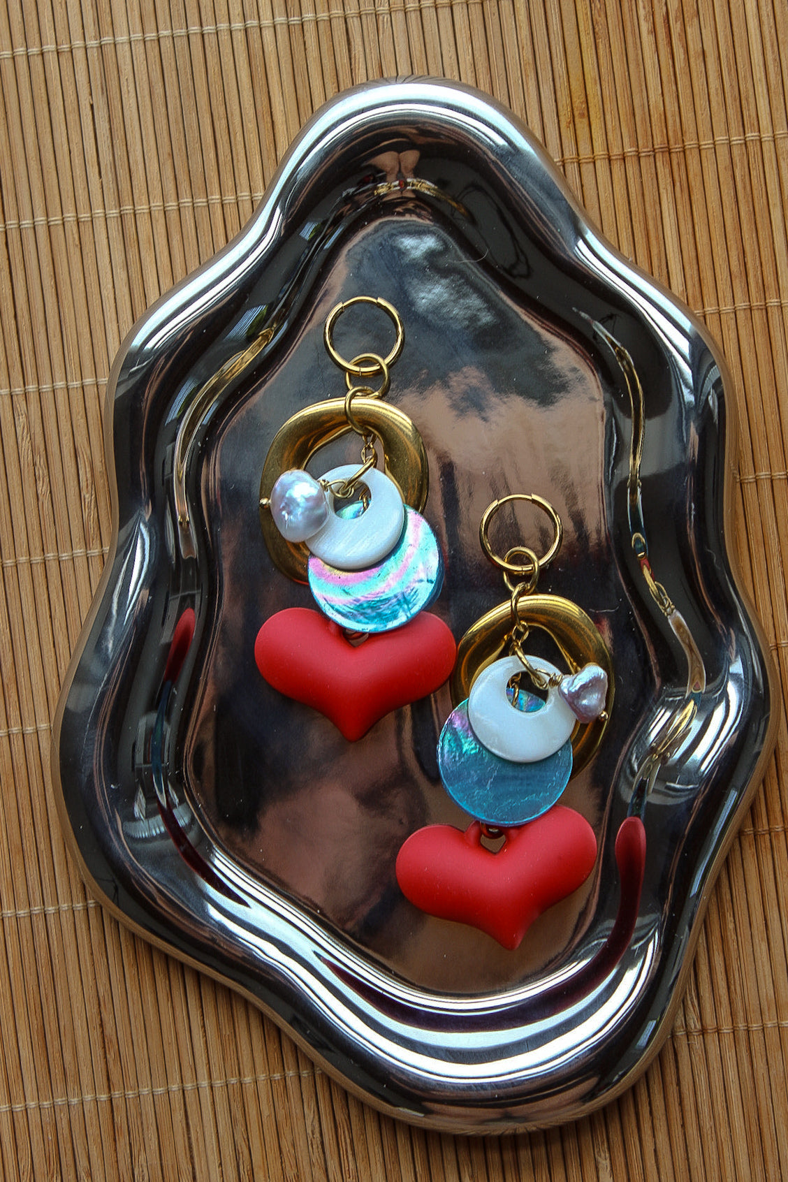 A pair of handmade dangle earrings displayed on a reflective silver tray over a woven bamboo mat. The earrings feature gold-tone rings, white pearl-like beads, iridescent blue beads, and red heart charms. Un par de pantallas colgantes artesanales sobre una bandeja plateada reflectante y un mantel de bambú tejido. El diseño incluye aros dorados, cuentas tipo perla blancas, cuentas azules iridiscentes y dijes de corazones rojos.