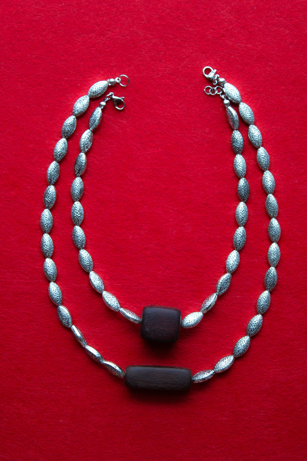 A silver-toned beaded necklace with textured oval beads and two dark rectangular center accents, displayed on a solid red background.