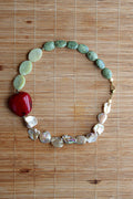 A handmade beaded necklace featuring a mix of green stone beads, flat pale green discs, a prominent glossy red central bead, and iridescent white shell beads, displayed on a brown bamboo mat.