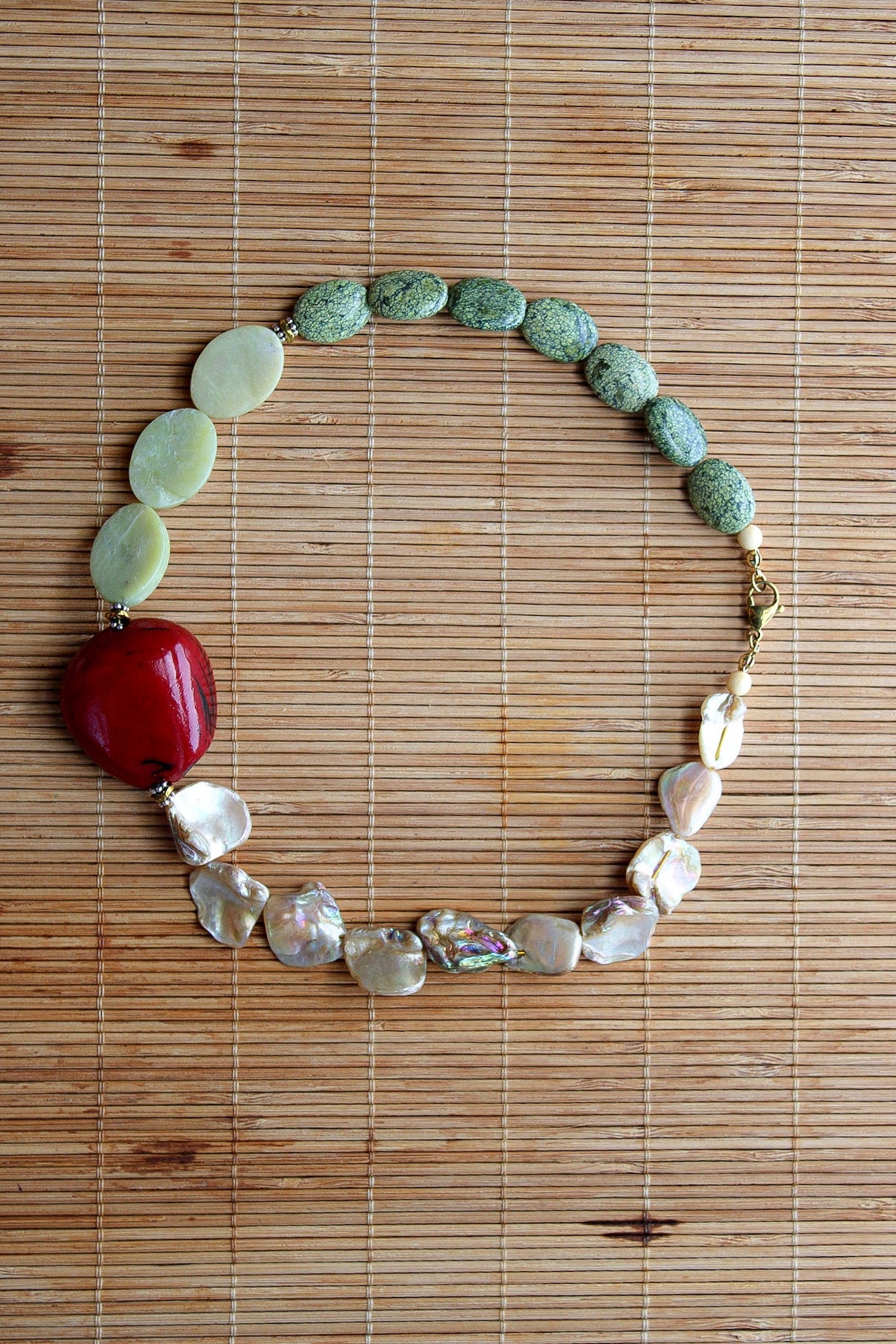 A handmade beaded necklace featuring a mix of green stone beads, flat pale green discs, a prominent glossy red central bead, and iridescent white shell beads, displayed on a brown bamboo mat.
