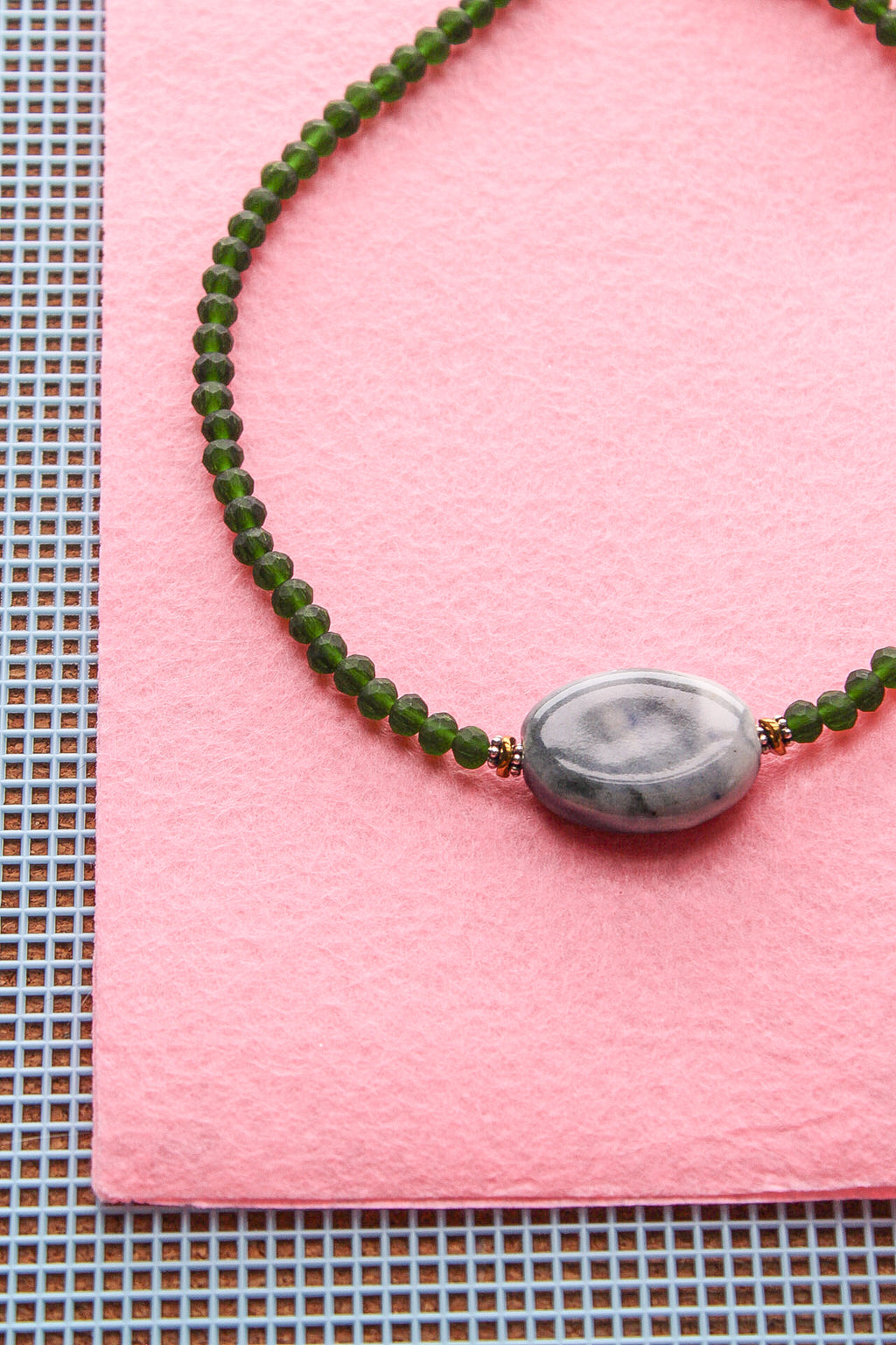 A beaded necklace with dark green faceted beads and a central gray marbled oval stone, displayed on a pink textured paper background.