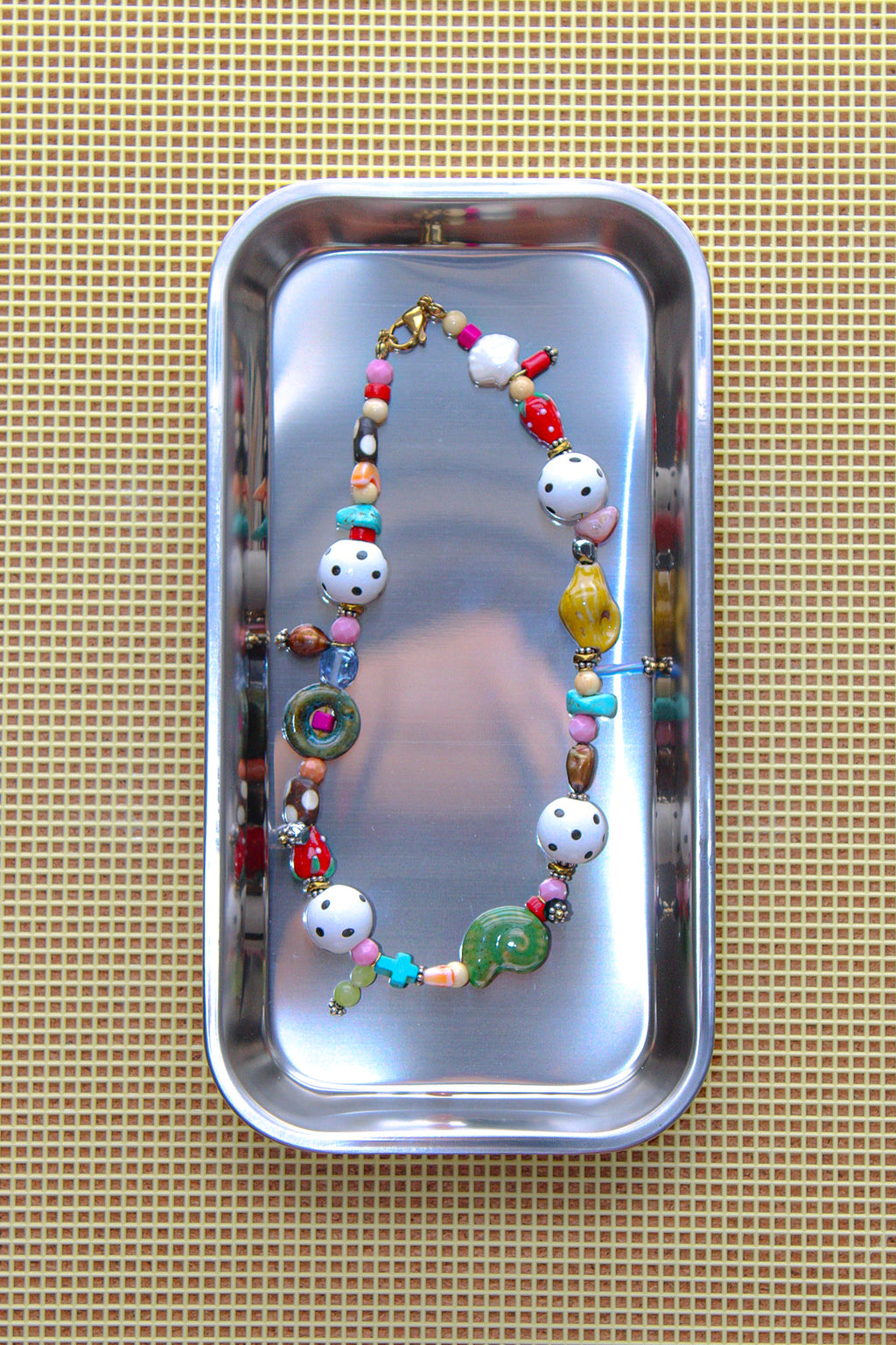A colorful beaded necklace with white polka-dot beads, green and yellow accents, and a gold clasp, displayed inside a rectangular silver metal tray on a beige woven mat.