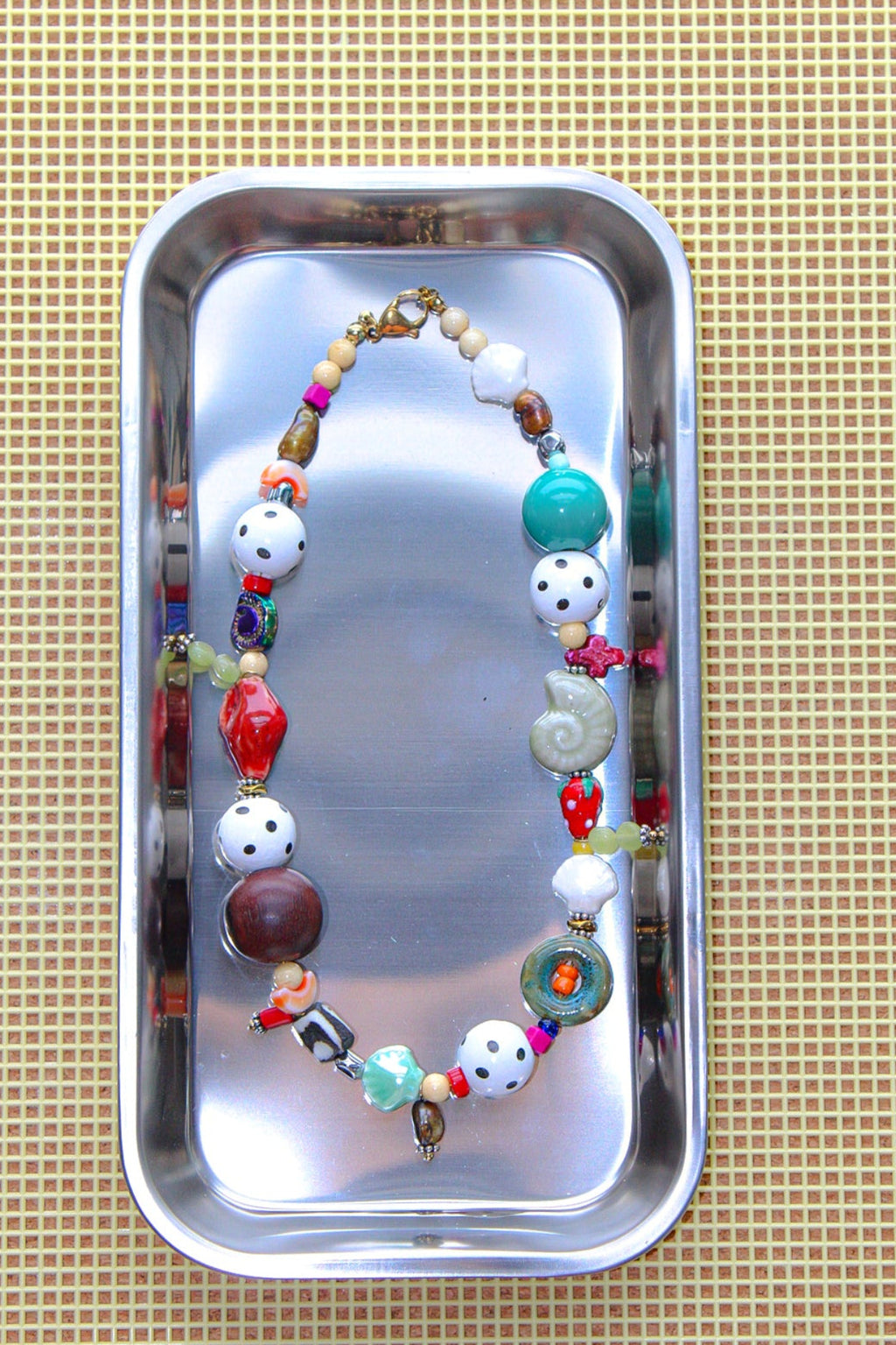 A colorful beaded necklace with white, red, green, and brown beads, displayed inside a small silver metal tray on a beige textured mat.