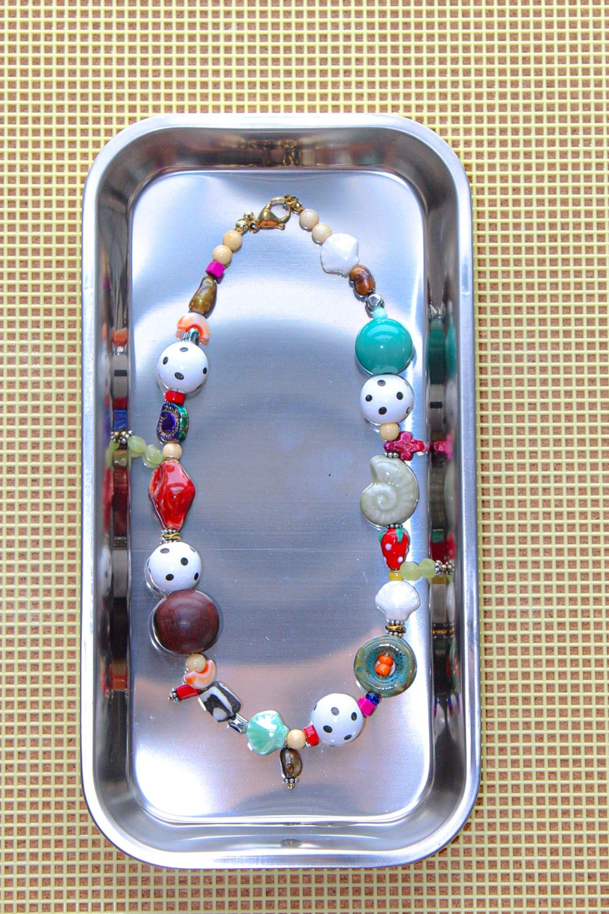 A colorful beaded necklace with white, red, green, and brown beads, displayed inside a small silver metal tray on a beige textured mat.