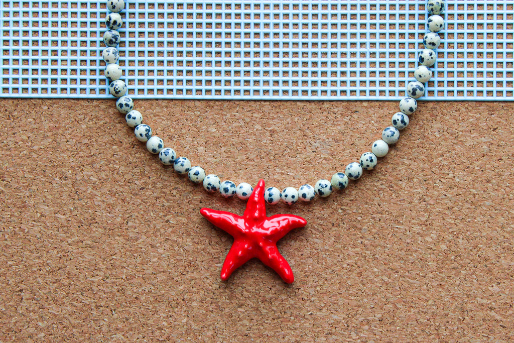 A beaded necklace with white beads featuring black speckles and a glossy red starfish pendant, displayed on a brown cork board with a blue grid mat in the background.
