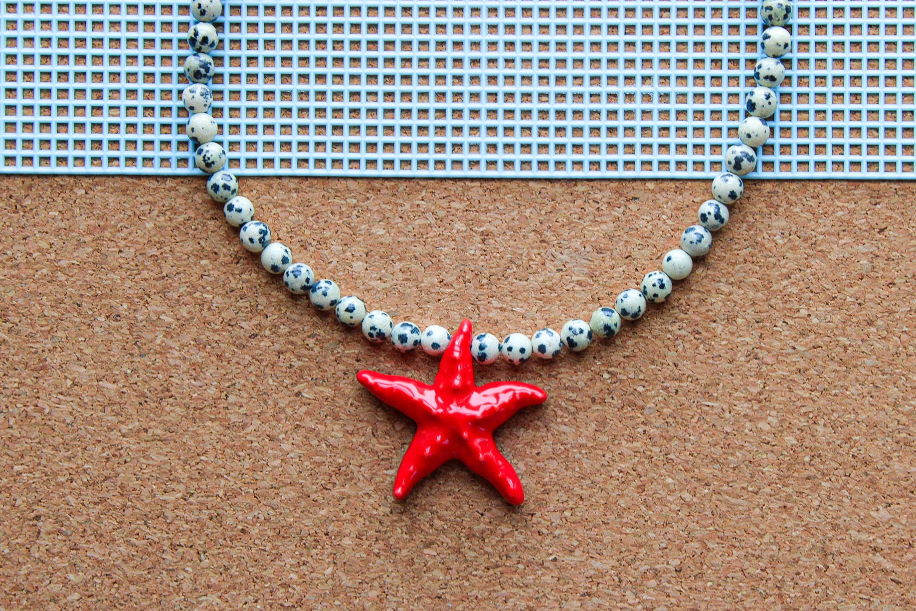 A beaded necklace with white beads featuring black speckles and a glossy red starfish pendant, displayed on a brown cork board with a blue grid mat in the background.