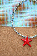 A beaded necklace with white and black speckled beads and a bright red starfish pendant, displayed on a cork board with a blue grid background.