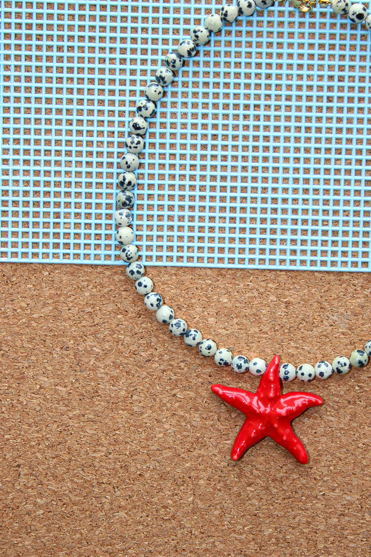 A beaded necklace with white and black speckled beads and a bright red starfish pendant, displayed on a cork board with a blue grid background.