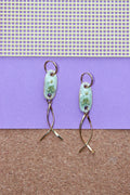 A pair of handmade dangle earrings with light blue-green floral enamel oval pendants and twisted gold-tone wire drops, displayed against a split purple and brown background. Pantallas de flores, aretes de flores, joyeria de ceramica, joyeria dorada, colgante.