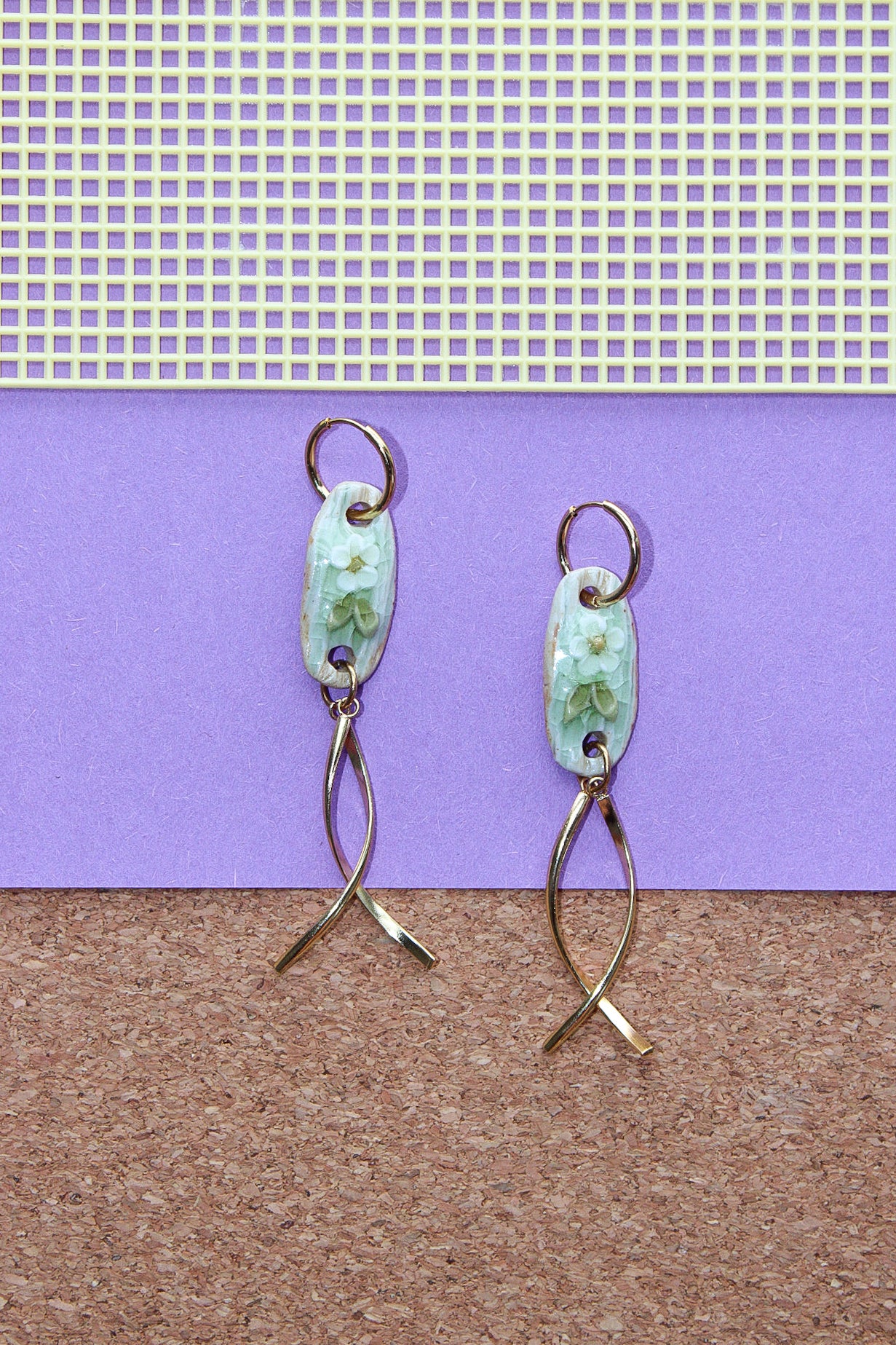 A pair of handmade dangle earrings with light blue-green floral enamel oval pendants and twisted gold-tone wire drops, displayed against a split purple and brown background. Pantallas de flores, aretes de flores, joyeria de ceramica, joyeria dorada, colgante.