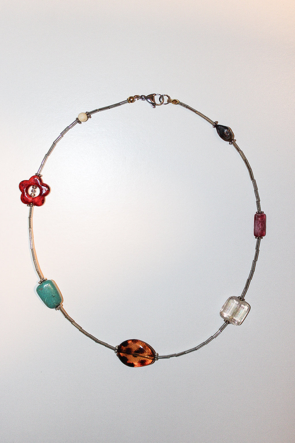 A delicate beaded necklace with a silver-toned wire base, featuring multiple colorful glass beads in red, turquoise, brown, and clear, displayed against a light gray background.