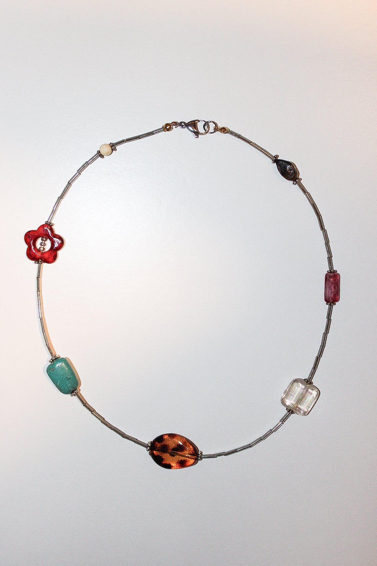 A delicate beaded necklace with a silver-toned wire base, featuring multiple colorful glass beads in red, turquoise, brown, and clear, displayed against a light gray background.