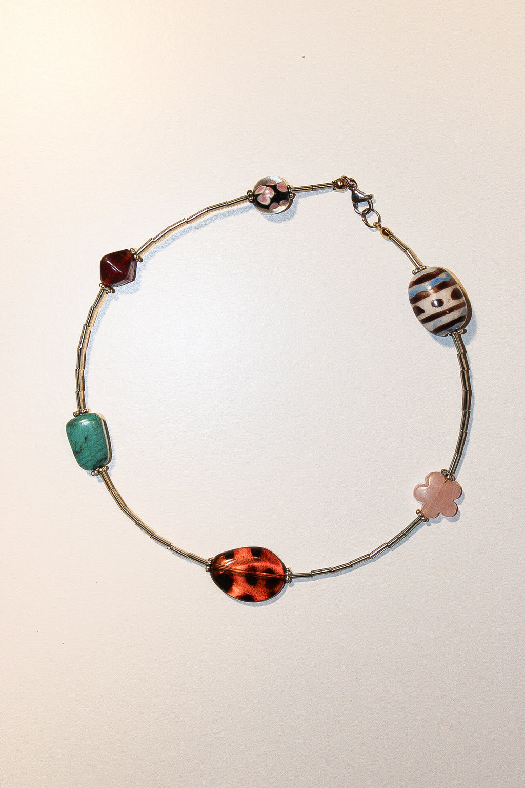A silver-toned beaded necklace with five distinct decorative beads, including a turquoise rectangular bead, a red faceted bead, a striped striped bead, a pink flower-shaped bead, and a red and black marbled bead, displayed on a plain off-white background.