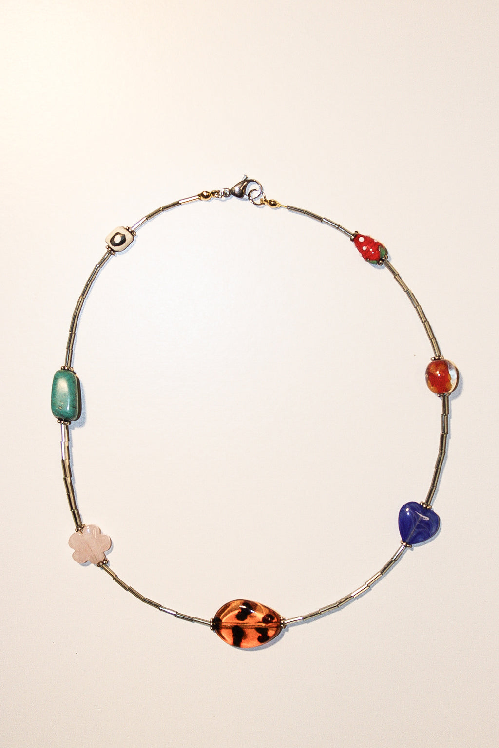 A delicate silver-toned beaded necklace featuring multiple colorful, organic-shaped beads in green, red, blue, orange, and pale pink, displayed on a plain off-white background.