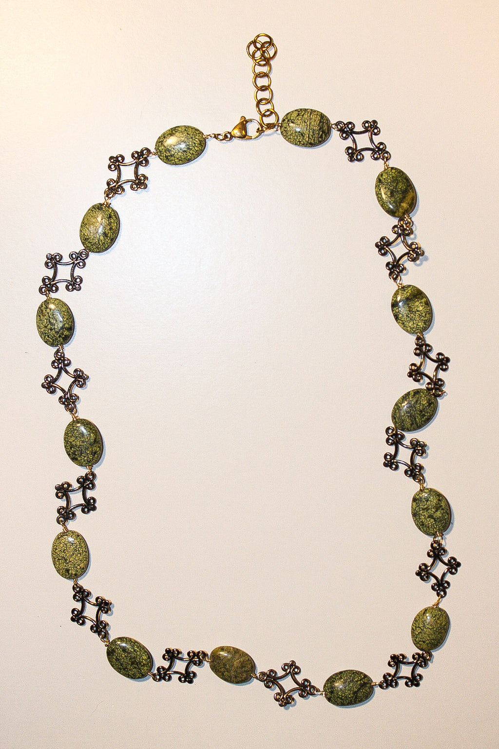 A vintage-style necklace featuring oval green marbled stones connected by dark bronze cross motifs and a gold-tone clasp, displayed on a plain light beige background.