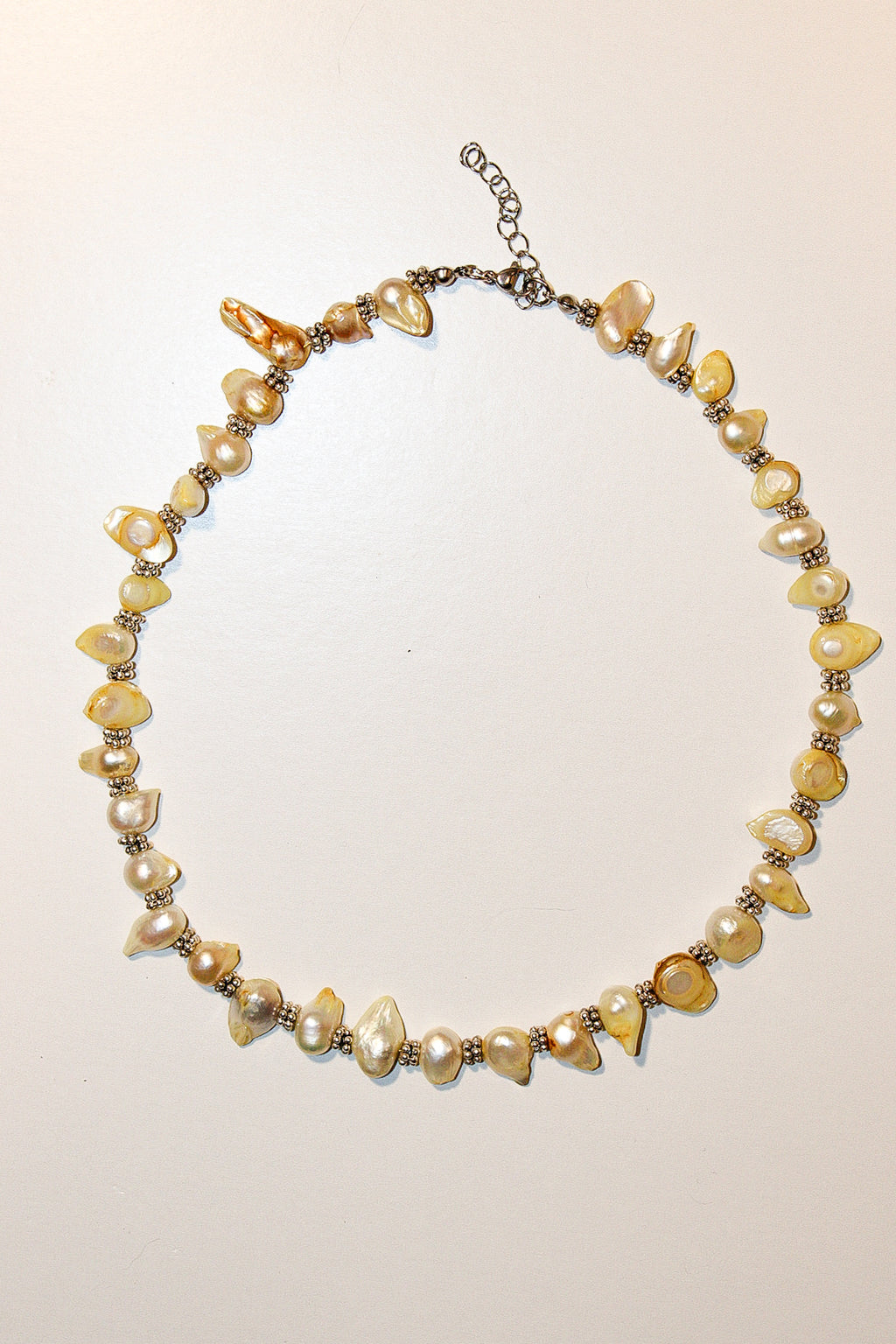 A handmade beaded necklace featuring irregular cream and yellow faux pearls, polished amber-like beads, and small metallic spacer beads on a silver-tone chain with an extender clasp, displayed on a plain off-white background.
