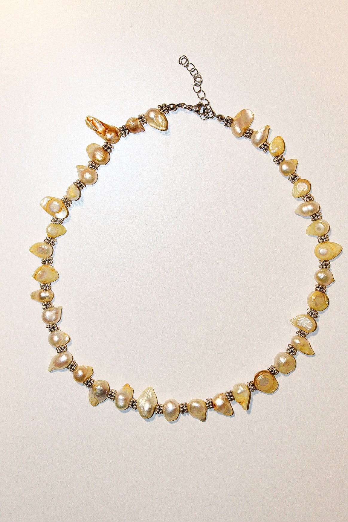 A handmade beaded necklace featuring irregular cream and yellow faux pearls, polished amber-like beads, and small metallic spacer beads on a silver-tone chain with an extender clasp, displayed on a plain off-white background.