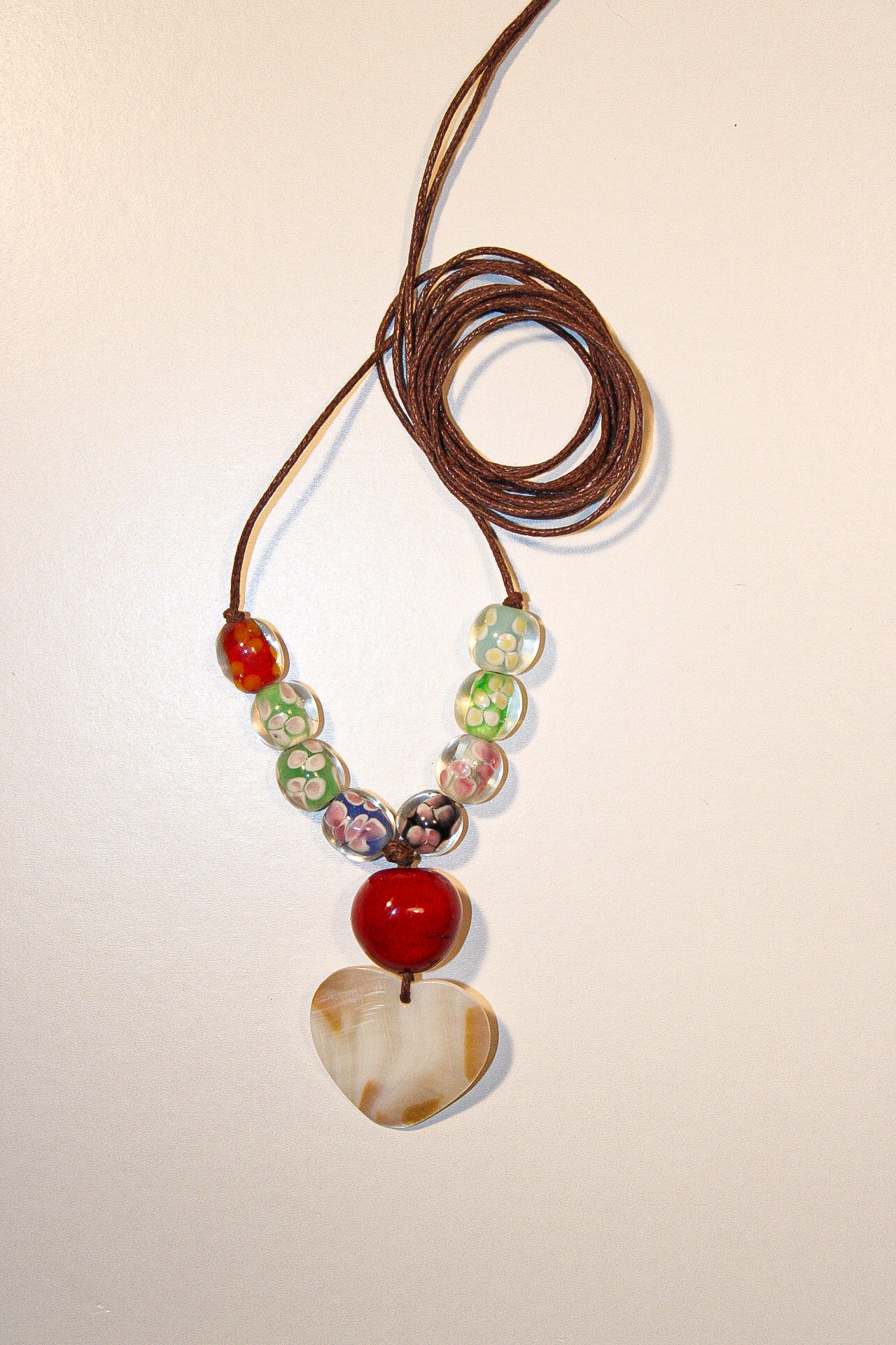 A handmade beaded necklace with a brown cord, featuring colorful patterned beads, a red bead, and a heart-shaped shell pendant, displayed on a plain off-white background.