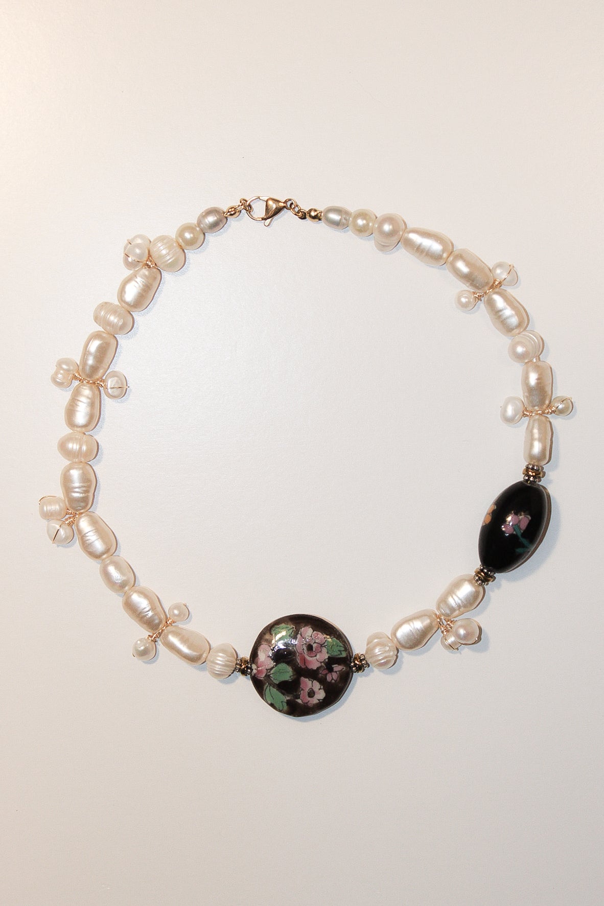 A pearl and glass bead necklace featuring a large oval black bead with a pink floral design, a matching round floral glass bead, and small gold-toned accents, displayed on a light beige background. Handmade necklaces by Marveyra handcrafted in Puerto Rico featuring unique artisan designs, stainless steel details, pearls, natural-inspired elements, charms, chains, and contemporary accents. Every piece is created to stand out with an alternative, beach-inspired, unisex aesthetic and can be layered or styled f