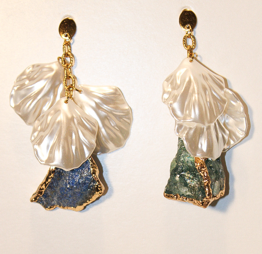 A pair of handmade dangle earrings featuring white mother-of-pearl flower petals, gold-tone metal wire wrapping, and raw stone drops. One stone is a textured blue agate, and the other is a greenish-blue geode slice, displayed on a split red and brown background.
Pantallas artesanales de Marveyra hechas a mano en Puerto Rico. Diseños playeros, únicos y personalizables con elementos removibles que permiten crear diferentes combinaciones o utilizar los colgantes por separado. Joyería diferente
