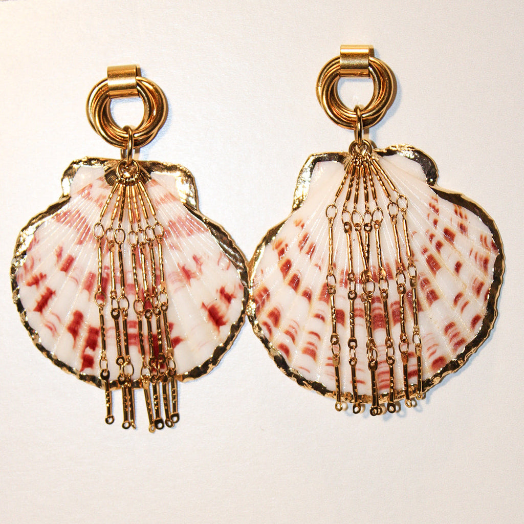 A pair of gold-tone statement earrings featuring scallop-shaped shells with pink and white marbled patterns and dangling beaded chains, set against a plain off-white background.A scallop shell pendant with red and white stripes, decorated with gold-toned chains and a gold clasp, displayed on a blue denim fabric background. Par de pantallas de concha caracol con cadenas colgantes doradas, grades y unicas, estilo llamativo. Pantallas playeras y tropicales a la moda.