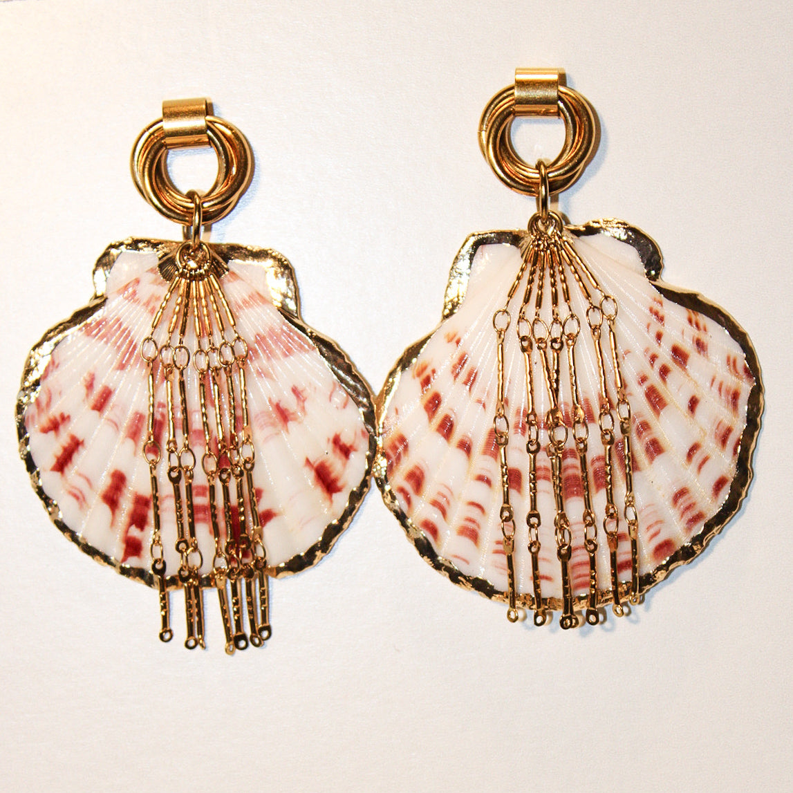 A pair of gold-tone statement earrings featuring scallop-shaped shells with pink and white marbled patterns and dangling beaded chains, set against a plain off-white background.A scallop shell pendant with red and white stripes, decorated with gold-toned chains and a gold clasp, displayed on a blue denim fabric background. Par de pantallas de concha caracol con cadenas colgantes doradas, grades y unicas, estilo llamativo. Pantallas playeras y tropicales a la moda.