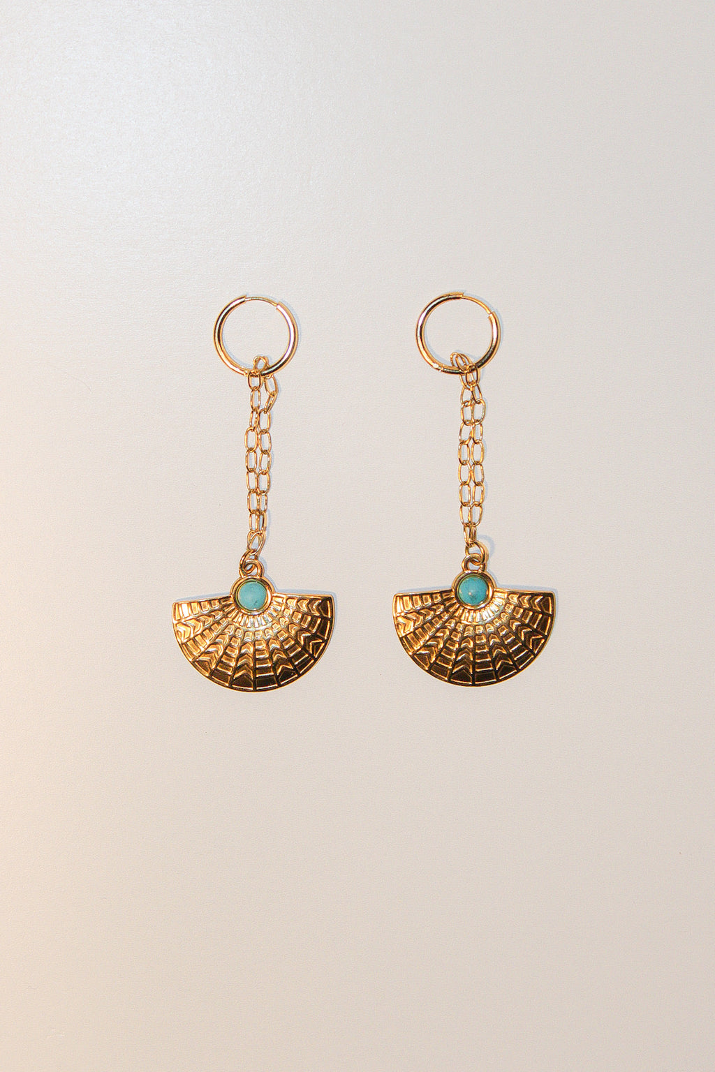 A pair of gold-toned dangle earrings with a textured fan-shaped drop and a small round turquoise stone at the center, shown on a plain light beige background.
Pantallas artesanales de Marveyra, hechas a mano en Puerto Rico con materiales resistentes. Este diseño de joyería alternativa y unisex cuenta con elementos removibles que permiten personalizar la pieza o utilizar los colgantes por separado como collar. Accesorios originales creados para quienes buscan destacar con un estilo diferente.