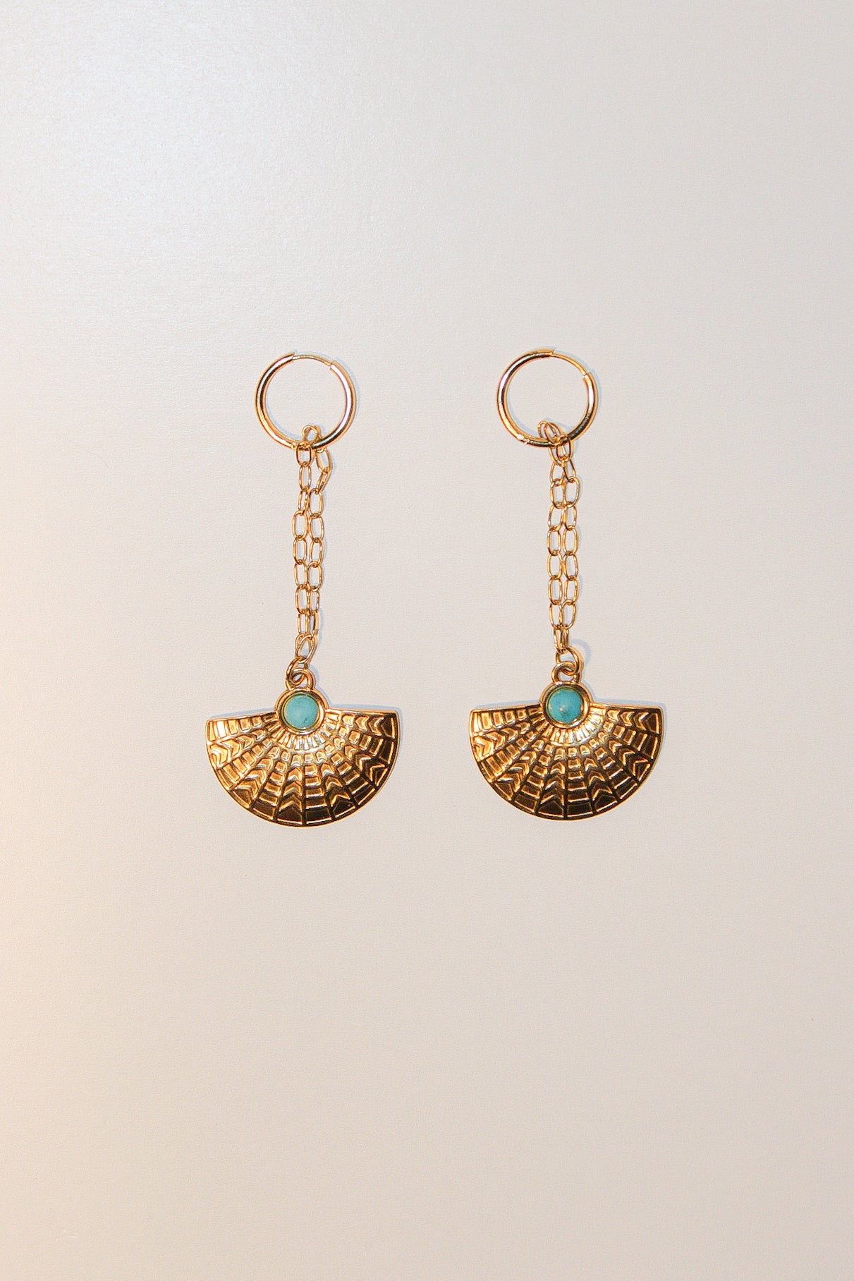 A pair of gold-toned dangle earrings with a textured fan-shaped drop and a small round turquoise stone at the center, shown on a plain light beige background.
Pantallas artesanales de Marveyra, hechas a mano en Puerto Rico con materiales resistentes. Este diseño de joyería alternativa y unisex cuenta con elementos removibles que permiten personalizar la pieza o utilizar los colgantes por separado como collar. Accesorios originales creados para quienes buscan destacar con un estilo diferente.