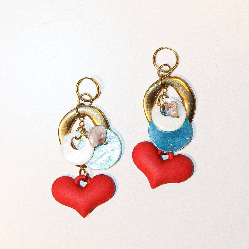 A pair of handmade dangle earrings displayed on a reflective silver tray over a woven bamboo mat. The earrings feature gold-tone rings, white pearl-like beads, iridescent blue beads, and red heart charms. Un par de pantallas colgantes artesanales sobre una bandeja plateada reflectante y un mantel de bambú tejido. El diseño incluye aros dorados, cuentas tipo perla blancas, cuentas azules iridiscentes y dijes de corazones rojos.