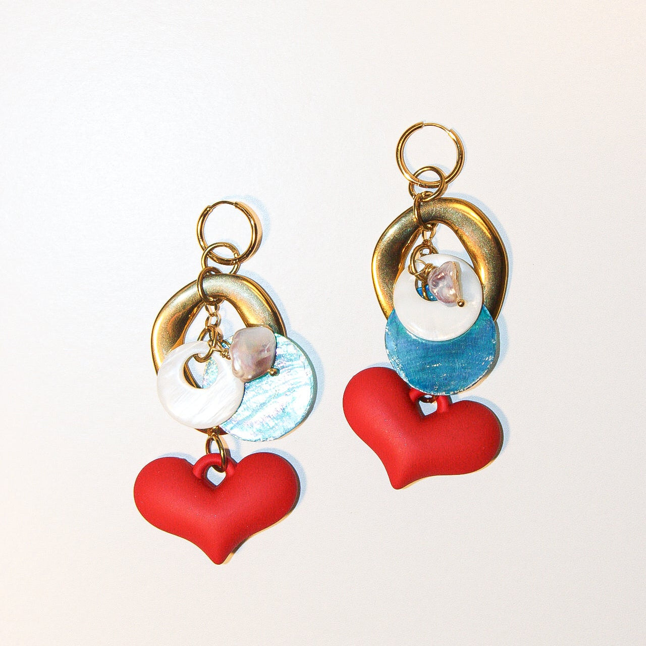 A pair of handmade dangle earrings displayed on a reflective silver tray over a woven bamboo mat. The earrings feature gold-tone rings, white pearl-like beads, iridescent blue beads, and red heart charms. Un par de pantallas colgantes artesanales sobre una bandeja plateada reflectante y un mantel de bambú tejido. El diseño incluye aros dorados, cuentas tipo perla blancas, cuentas azules iridiscentes y dijes de corazones rojos.