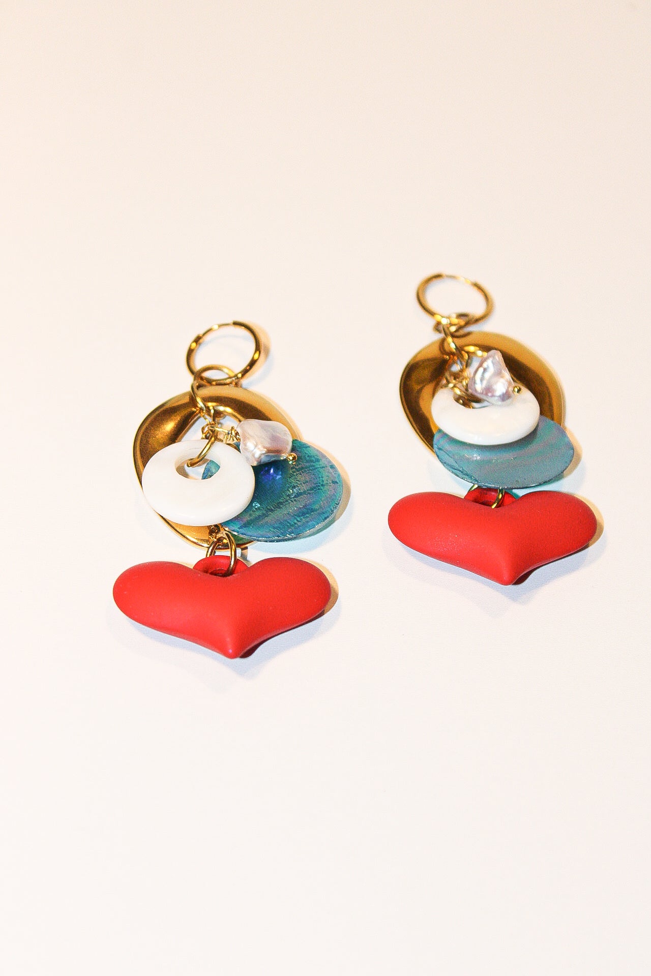 A pair of handmade dangle earrings displayed on a reflective silver tray over a woven bamboo mat. The earrings feature gold-tone rings, white pearl-like beads, iridescent blue beads, and red heart charms. Un par de pantallas colgantes artesanales sobre una bandeja plateada reflectante y un mantel de bambú tejido. El diseño incluye aros dorados, cuentas tipo perla blancas, cuentas azules iridiscentes y dijes de corazones rojos.