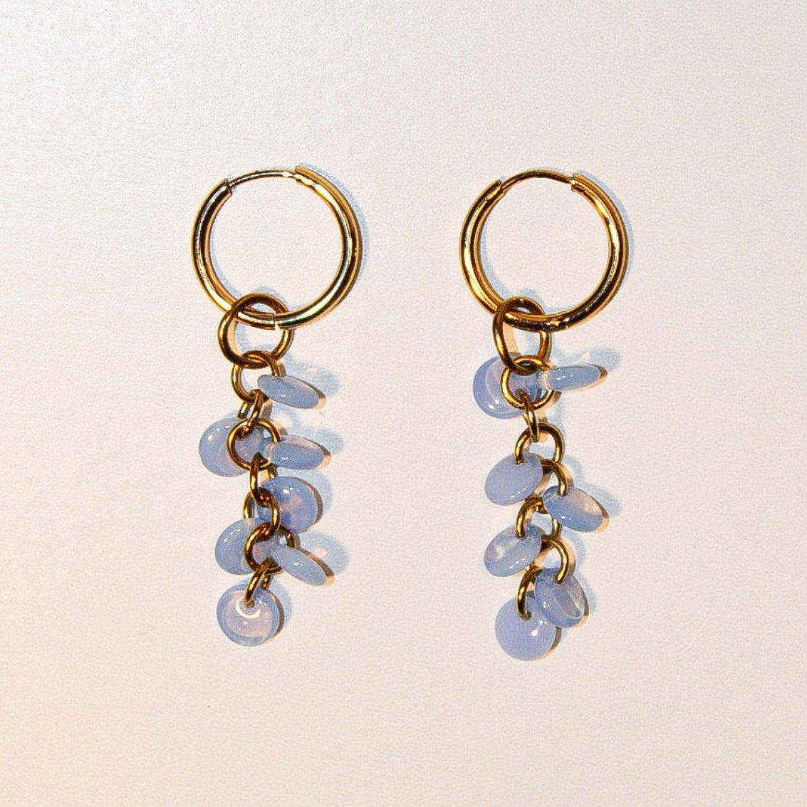 A pair of gold hoop dangle earrings with small light blue faceted beads, displayed on a plain beige background. Pantallas colgantes con gotas de cristales azules colgando. Pantallas delicadas y sencillez. A la moda, estilo único.