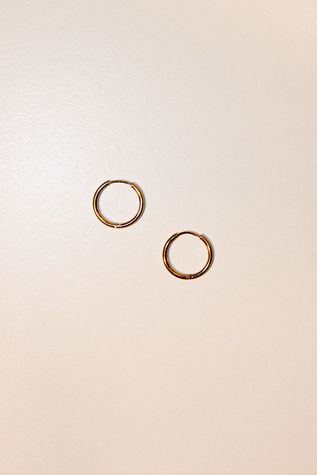 A pair of thin gold hoop earrings displayed on a plain, warm off-white background.