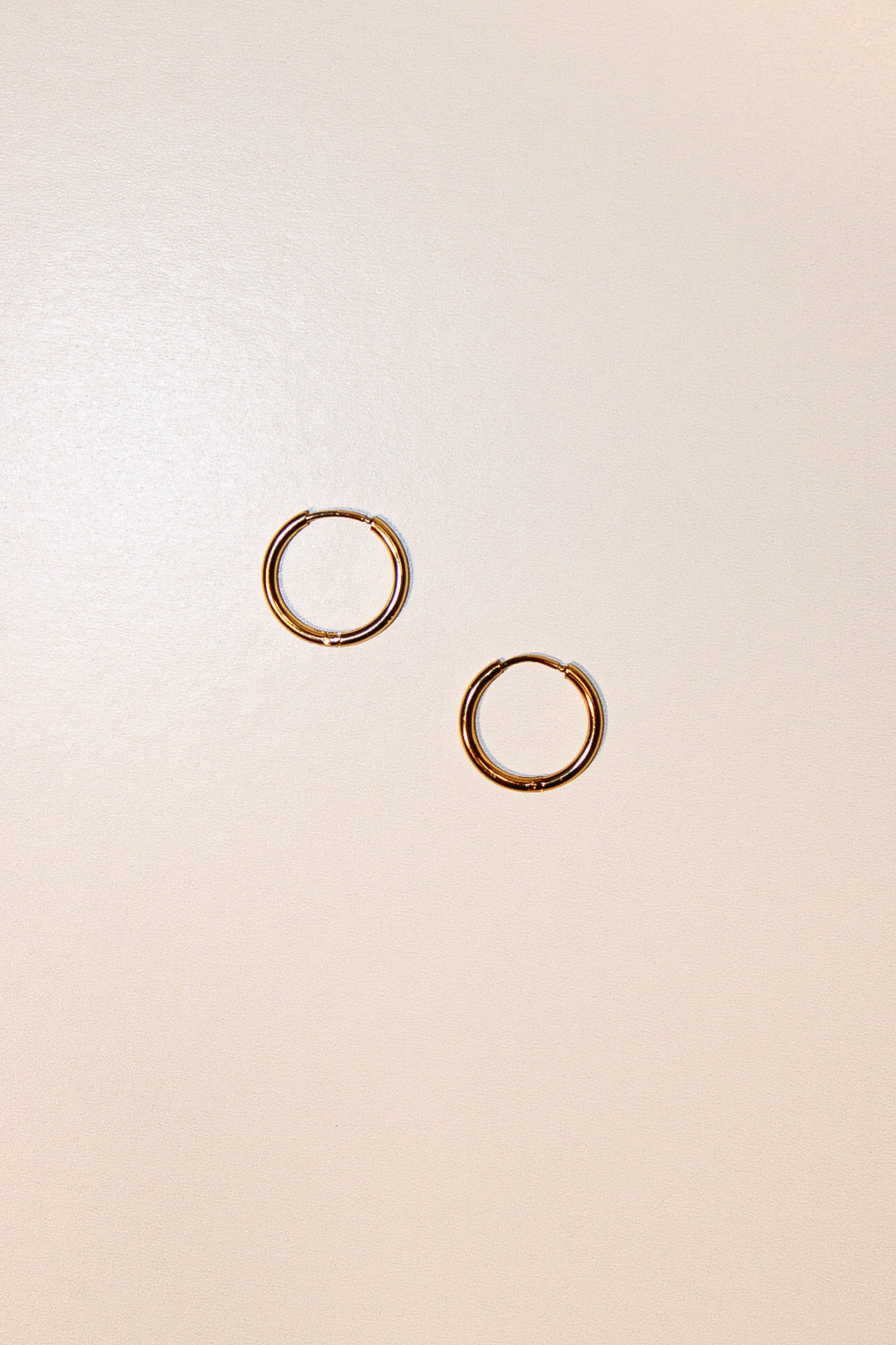A pair of thin gold hoop earrings displayed on a plain, warm off-white background.