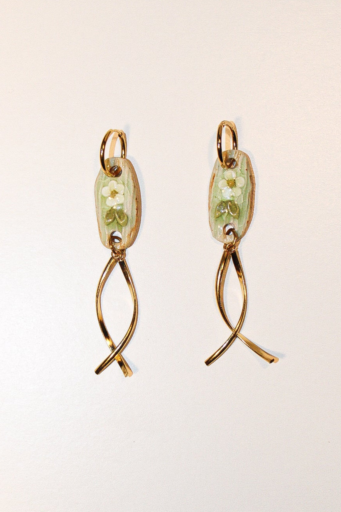 A pair of handmade dangle earrings with light blue-green floral enamel oval pendants and twisted gold-tone wire drops, displayed against a split purple and brown background. Pantallas de flores, aretes de flores, joyeria de ceramica, joyeria dorada, colgante.