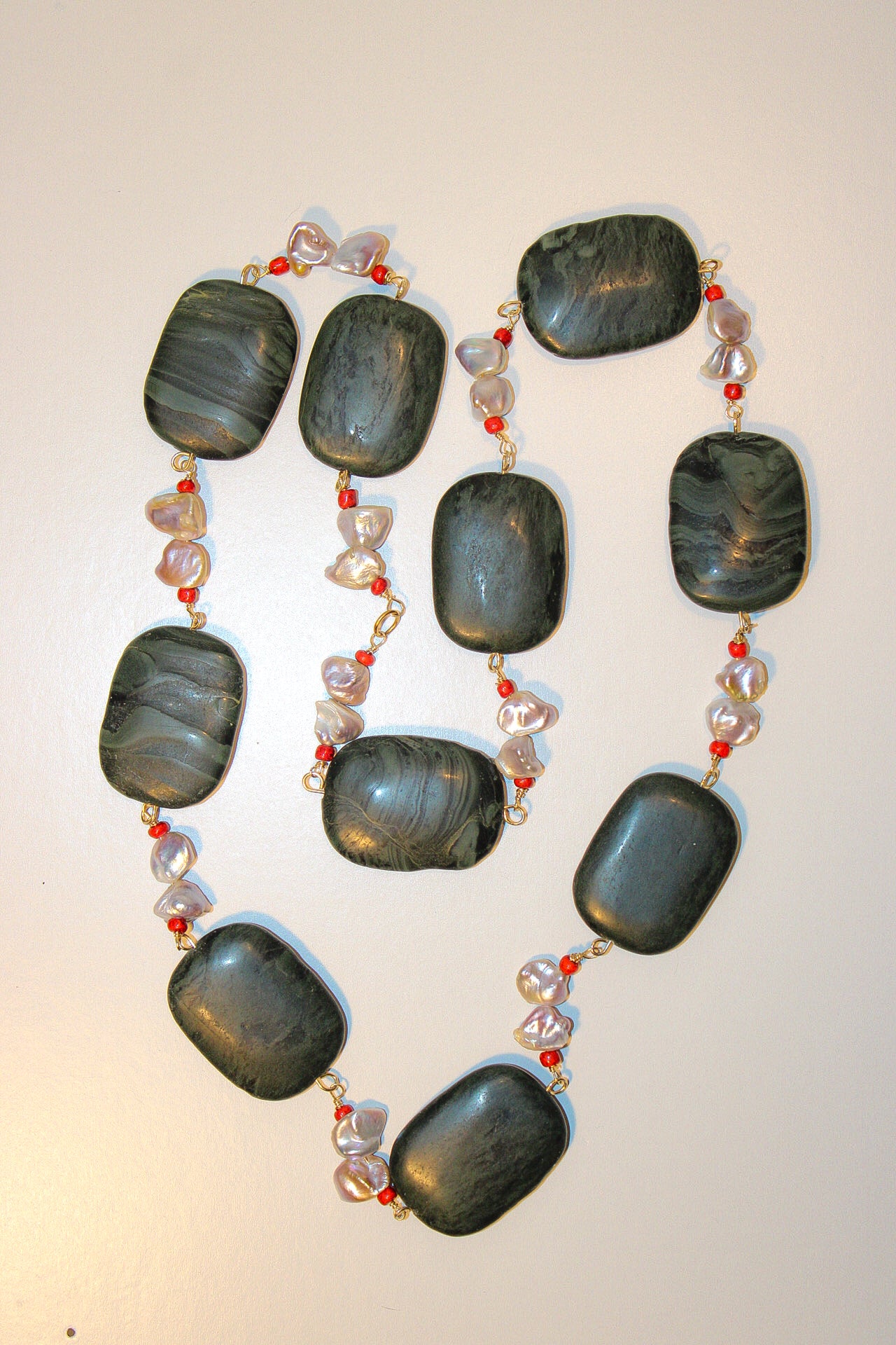 A beaded necklace featuring large, dark gray polished stone oval beads separated by small strands of clear faceted beads and red seed beads on a gold-tone chain, displayed on a plain off-white background.