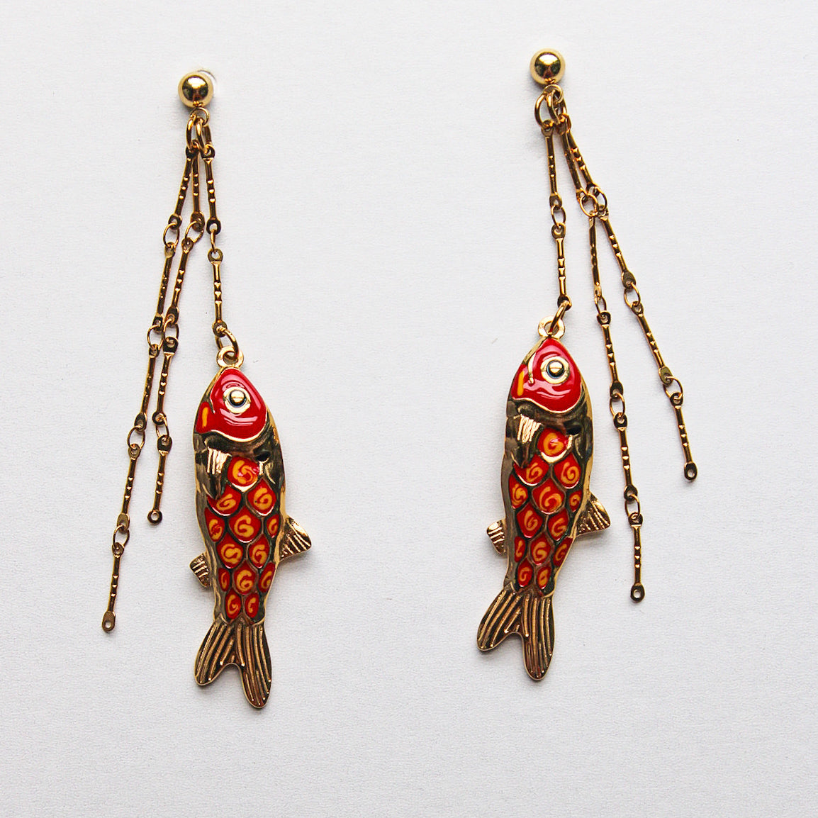 A single dangling fish-shaped earring in gold-tone metal with red and orange enamel detailing, photographed on a blue grid-patterned background. Pantallas artesanales de Marveyra, hechas a mano en Puerto Rico con materiales resistentes. Este diseño de joyería alternativa y unisex cuenta con elementos removibles que permiten personalizar la pieza o utilizar los colgantes por separado como collar. Accesorios originales creados para quienes buscan destacar con un estilo diferente. Pantallas de peces.