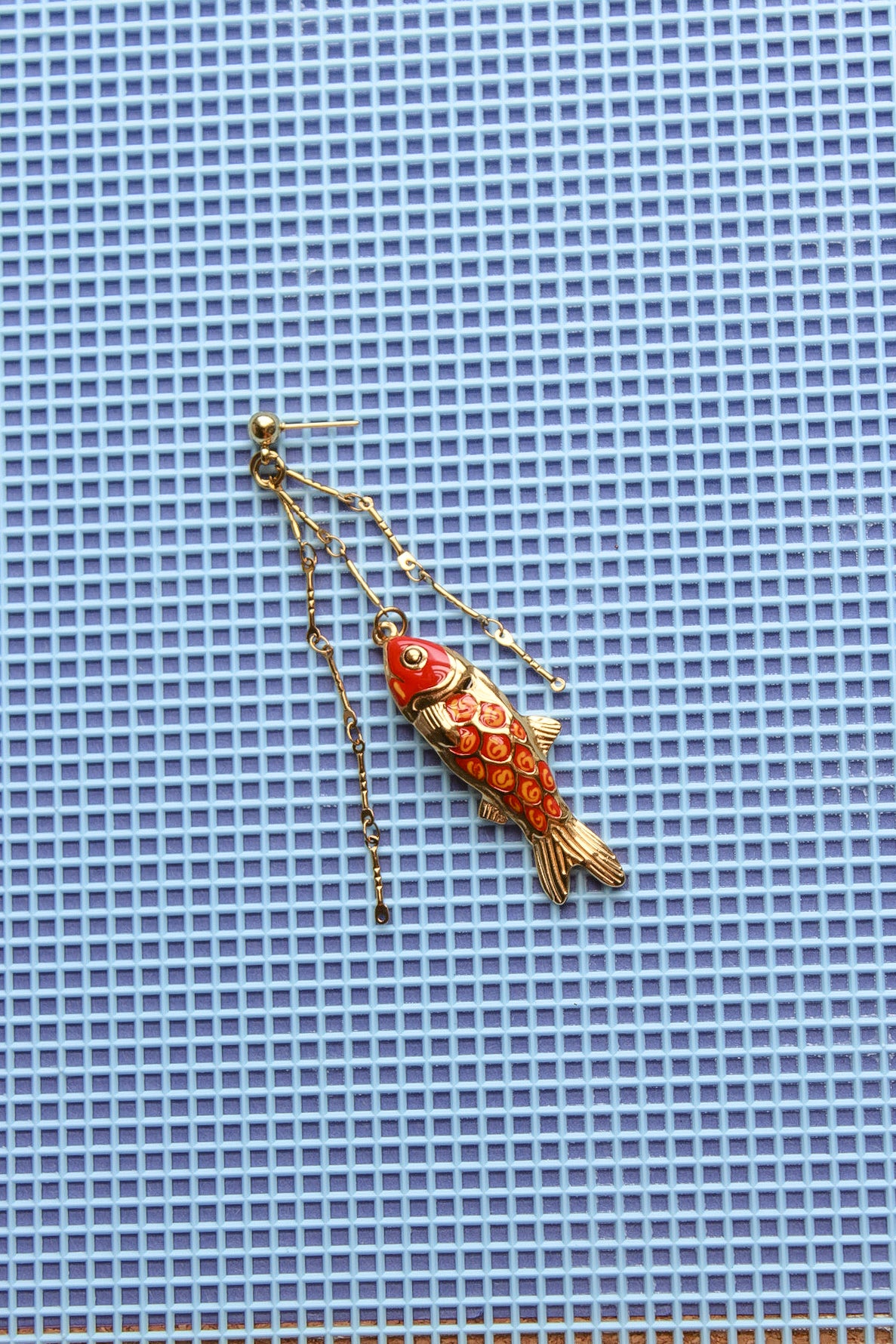 A single dangling fish-shaped earring in gold-tone metal with red and orange enamel detailing, photographed on a blue grid-patterned background. Pantallas artesanales de Marveyra, hechas a mano en Puerto Rico con materiales resistentes. Este diseño de joyería alternativa y unisex cuenta con elementos removibles que permiten personalizar la pieza o utilizar los colgantes por separado como collar. Accesorios originales creados para quienes buscan destacar con un estilo diferente. Pantallas de peces.