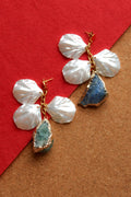 A pair of handmade dangle earrings featuring white mother-of-pearl flower petals, gold-tone metal wire wrapping, and raw stone drops. One stone is a textured blue agate, and the other is a greenish-blue geode slice, displayed on a split red and brown background.
Pantallas artesanales de Marveyra hechas a mano en Puerto Rico. Diseños playeros, únicos y personalizables con elementos removibles que permiten crear diferentes combinaciones o utilizar los colgantes por separado. Joyería diferente